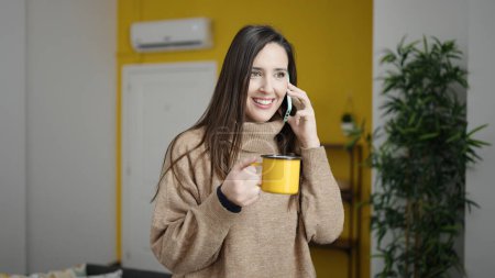Photo for Beautiful hispanic woman speaking on the phone drinking a cup of coffee at home - Royalty Free Image