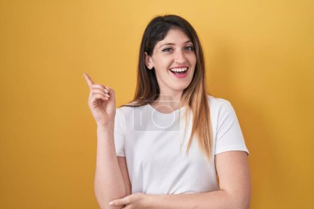 Photo for Young brunette woman standing over yellow background with a big smile on face, pointing with hand and finger to the side looking at the camera. - Royalty Free Image