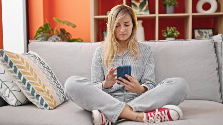 Téléchargez les photos : Jeune femme blonde écoutant de la musique assise sur le canapé à la maison - en image libre de droit