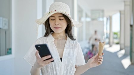Téléchargez les photos : Jeune femme chinoise touriste manger de la crème glacée en utilisant smartphone à la rue - en image libre de droit