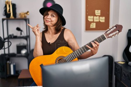 Photo for Middle age hispanic woman playing classic guitar at music studio smiling happy pointing with hand and finger to the side - Royalty Free Image