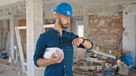 Photo for Young hispanic man architect wearing hardhat holding blueprints looking at wrist watch at construction site - Royalty Free Image