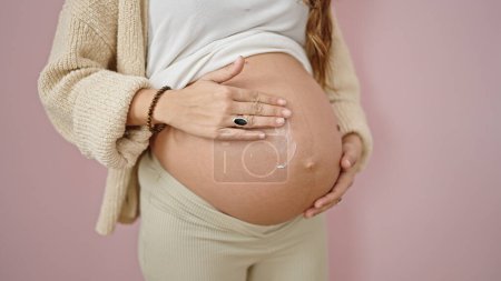 Photo for Young pregnant woman massaging belly applying skin care lotion over isolated pink background - Royalty Free Image