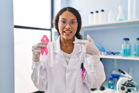 Photo for Young hispanic doctor woman working at scientist laboratory holding pink ribbon smiling happy pointing with hand and finger - Royalty Free Image