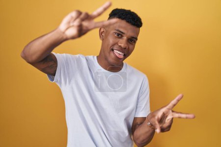 Photo for Young hispanic man standing over yellow background smiling with tongue out showing fingers of both hands doing victory sign. number two. - Royalty Free Image