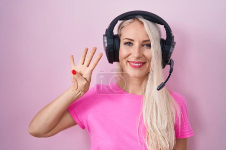 Téléchargez les photos : Femme caucasienne écoutant de la musique à l'aide d'écouteurs montrant et pointant vers le haut avec les doigts numéro quatre tout en souriant confiant et heureux. - en image libre de droit