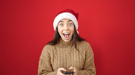 Foto de Joven mujer hispana hermosa usando sombrero de Navidad usando teléfono inteligente con expresión sorpresa sobre fondo rojo aislado - Imagen libre de derechos