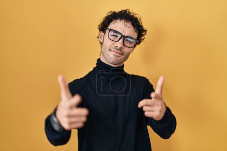 Foto de Hispanic man standing over yellow background pointing fingers to camera with happy and funny face. good energy and vibes. - Imagen libre de derechos