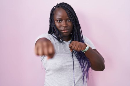 Foto de Mujer africana joven de pie sobre fondo rosa golpeando puño para luchar, ataque agresivo y enojado, amenaza y violencia - Imagen libre de derechos