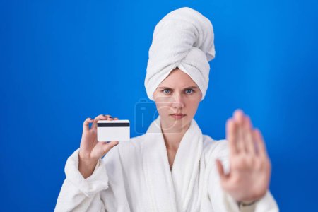 Foto de Mujer rubia caucásica con albornoz sosteniendo tarjeta de crédito con la mano abierta haciendo stop sign con expresión seria y segura, gesto de defensa - Imagen libre de derechos