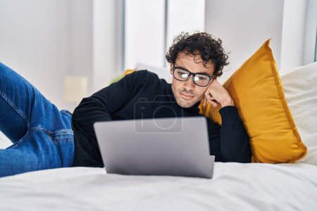Téléchargez les photos : Jeune homme hispanique utilisant un ordinateur portable couché sur le lit dans la chambre - en image libre de droit