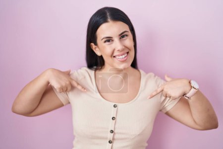 Foto de Mujer hispana joven de pie sobre fondo rosa mirando confiado con sonrisa en la cara, señalándose con los dedos orgullosos y felices. - Imagen libre de derechos
