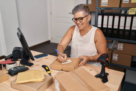 Téléchargez les photos : Femme d'âge moyen ecommerce travailleur d'affaires écrivant sur le paquet au bureau - en image libre de droit