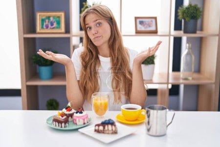 Photo for Young caucasian woman eating pastries t for breakfast clueless and confused expression with arms and hands raised. doubt concept. - Royalty Free Image