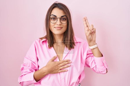 Foto de Mujer hispana joven usando gafas de pie sobre fondo rosa sonriendo jurando con la mano en el pecho y los dedos hacia arriba, haciendo un juramento de lealtad promesa - Imagen libre de derechos