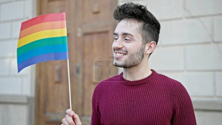 Foto de Joven hombre hispano sonriendo confiado sosteniendo bandera de arco iris en la calle - Imagen libre de derechos