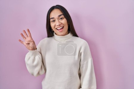 Foto de Joven mujer del sur asiático de pie sobre fondo rosa mostrando y señalando hacia arriba con los dedos número cuatro mientras sonríe confiado y feliz. - Imagen libre de derechos