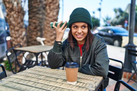 Photo for Young beautiful hispanic woman listening voice message by smartphone sitting on table at coffee shop terrace - Royalty Free Image