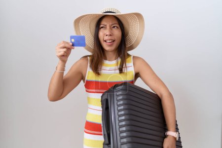 Foto de Mujer china de mediana edad sosteniendo maleta y tarjeta de crédito enojada y loca gritando frustrada y furiosa, gritando de rabia. rabia y concepto agresivo. - Imagen libre de derechos