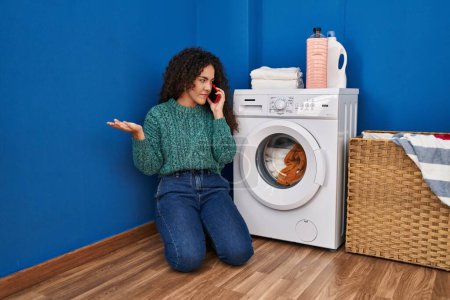 Téléchargez les photos : Jeune belle femme hispanique parlant sur smartphone avec technicien pour machine à laver cassée à la buanderie - en image libre de droit