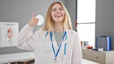 Foto de Joven rubia doctora haciendo pulgares arriba sonriendo en la clínica - Imagen libre de derechos