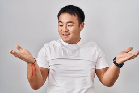 Téléchargez les photos : Jeune homme chinois debout sur fond blanc souriant montrant les paumes ouvertes des deux mains, présentant et comparant la publicité et l'équilibre - en image libre de droit