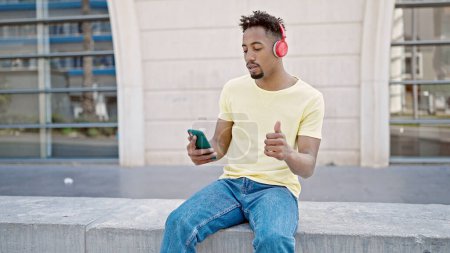 Photo for African american man listening to music using smartphone with thumb up at street - Royalty Free Image