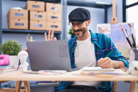 Foto de Hombre hispano con barba haciendo videollamada en línea en el estudio de arte haciendo signo bien con los dedos, sonriendo gesto amistoso excelente símbolo - Imagen libre de derechos