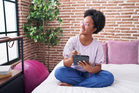 Téléchargez les photos : African american woman using touchpad sitting on bed at bedroom - en image libre de droit
