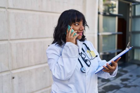 Téléchargez les photos : Young beautiful latin woman doctor talking on smartphone reading document at hospital - en image libre de droit
