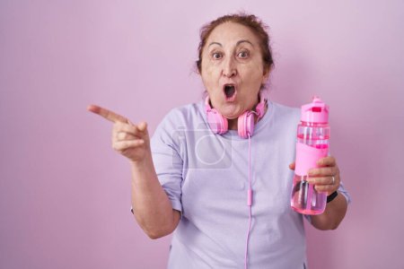 Foto de Mujer mayor con ropa deportiva y auriculares sorprendidos señalando con el dedo hacia un lado, boca abierta expresión asombrada. - Imagen libre de derechos