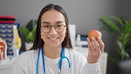 Photo for Young beautiful hispanic woman doctor smiling confident holding apple at clinic - Royalty Free Image