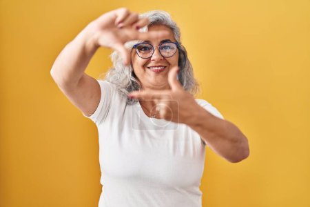 Téléchargez les photos : Femme d'âge moyen avec les cheveux gris debout sur fond jaune souriant faisant cadre avec les mains et les doigts avec le visage heureux. concept de créativité et photographie. - en image libre de droit