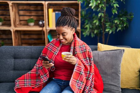 Foto de African american woman drinking coffee and using smartphone at home - Imagen libre de derechos
