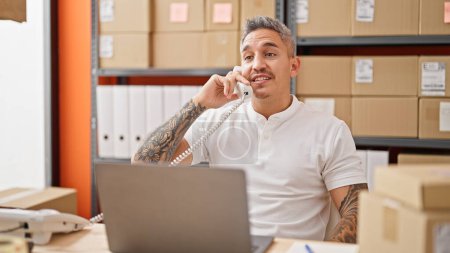 Téléchargez les photos : Jeune homme hispanique commerce électronique travailleur d'affaires en utilisant un ordinateur portable parlant au téléphone souriant au bureau - en image libre de droit
