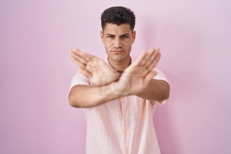 Téléchargez les photos : Young hispanic man standing over pink background rejection expression crossing arms and palms doing negative sign, angry face - en image libre de droit