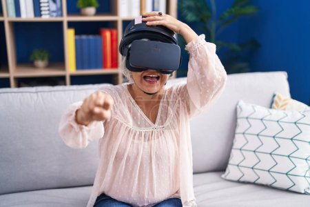 Téléchargez les photos : Femme aux cheveux gris moyen âge jouant à un jeu vidéo de moto en utilisant des lunettes de réalité virtuelle à la maison - en image libre de droit