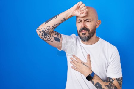 Foto de Hombre hispano con tatuajes de pie sobre fondo azul tocando la frente por enfermedad y fiebre, gripe y resfriado, virus enfermo - Imagen libre de derechos
