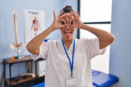 Foto de Una joven hispana que trabaja en una clínica de rehabilitación haciendo un buen gesto como binoculares sacando la lengua, ojos mirando a través de los dedos. expresión loca. - Imagen libre de derechos