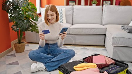 Téléchargez les photos : Jeune femme blonde shopping vol avec smartphone et carte de crédit assis sur le sol à la maison - en image libre de droit