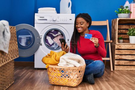 Foto de Mujer afroamericana usando teléfono inteligente y lavado de ropa con tarjeta de crédito en la lavandería - Imagen libre de derechos