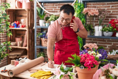 Photo for Young chinese man florist talking on smartphone writing on notebook at florist - Royalty Free Image