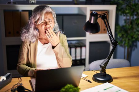 Téléchargez les photos : Femme d'âge moyen avec les cheveux gris travaillant à l'aide d'un ordinateur portable tard dans la nuit ennuyé bâillement fatigué couvrant la bouche avec la main. agitation et somnolence. - en image libre de droit
