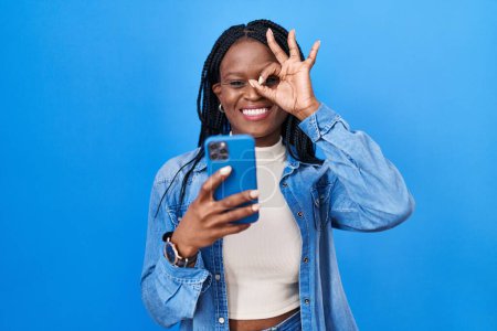 Téléchargez les photos : Femme africaine avec des tresses en utilisant le message de dactylographie smartphone souriant heureux faire ok signe avec la main sur les yeux regardant à travers les doigts - en image libre de droit