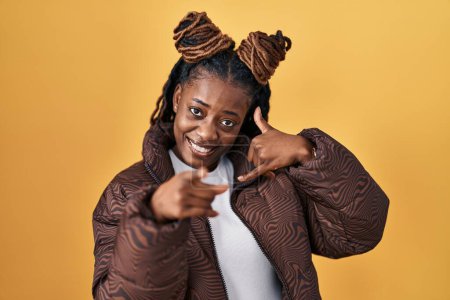 Téléchargez les photos : Femme africaine avec les cheveux tressés debout sur fond jaune souriant faire parler au téléphone geste et vous pointant du doigt. Appelle-moi. - en image libre de droit