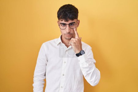 Téléchargez les photos : Jeune homme hispanique debout sur fond jaune pointant vers l'œil vous regardant geste, expression suspecte - en image libre de droit