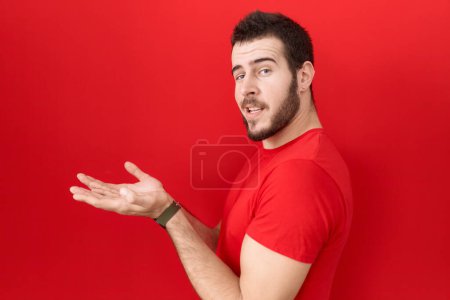Photo for Young hispanic man wearing casual red t shirt pointing aside with hands open palms showing copy space, presenting advertisement smiling excited happy - Royalty Free Image