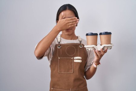 Photo for Young hispanic woman wearing professional waitress apron holding coffee covering eyes with hand, looking serious and sad. sightless, hiding and rejection concept - Royalty Free Image