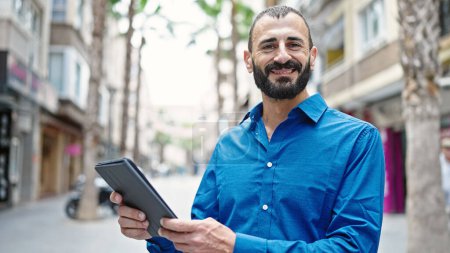 Téléchargez les photos : Jeune homme hispanique souriant confiant en utilisant touchpad à la rue - en image libre de droit