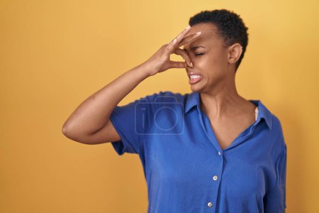 Téléchargez les photos : Femme afro-américaine debout sur fond jaune sentant quelque chose de puant et dégoûtant, odeur intolérable, retenant souffle avec les doigts sur le nez. mauvaise odeur - en image libre de droit